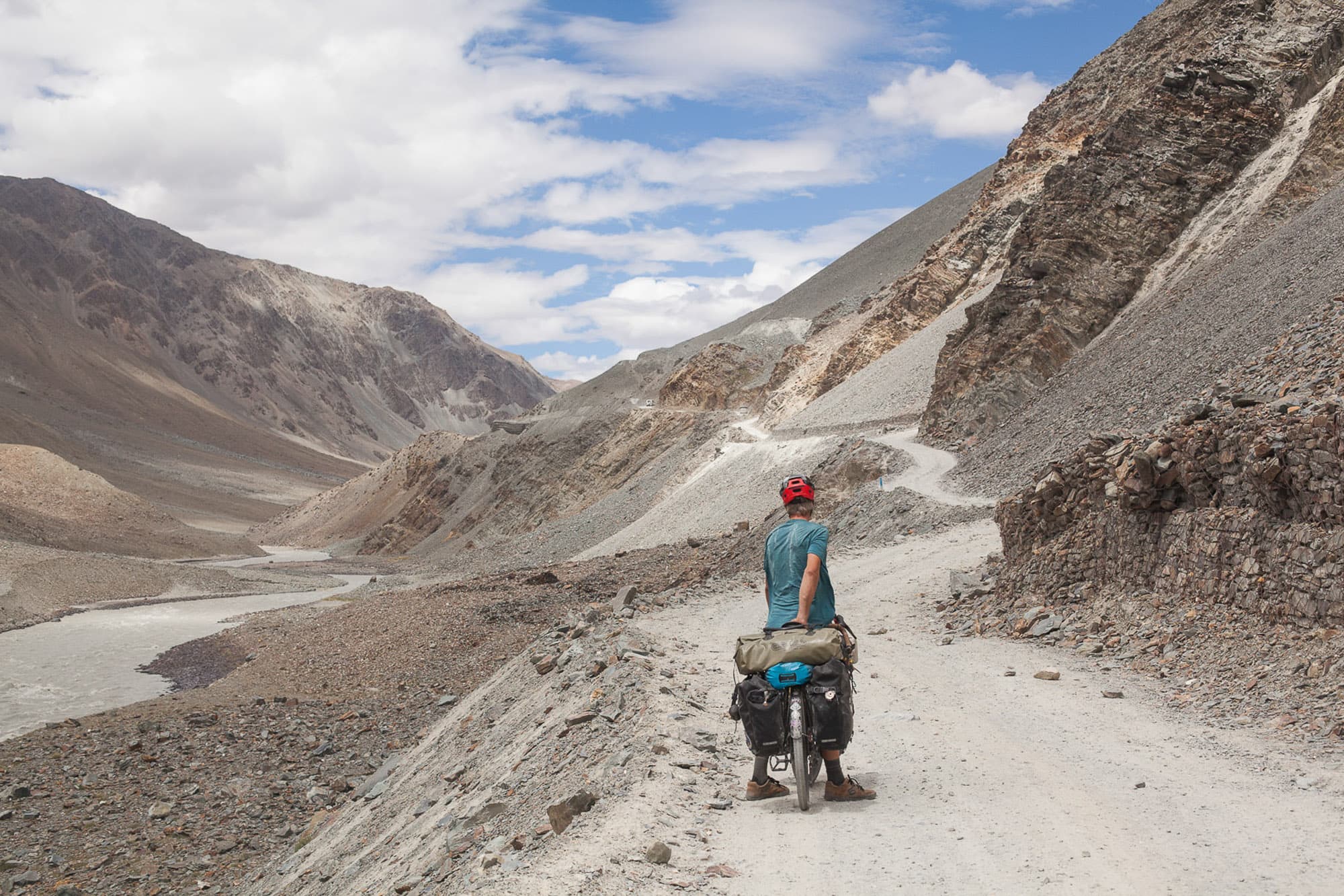 A cyclist with a loaded bike on a gravel road in a mountain valley.