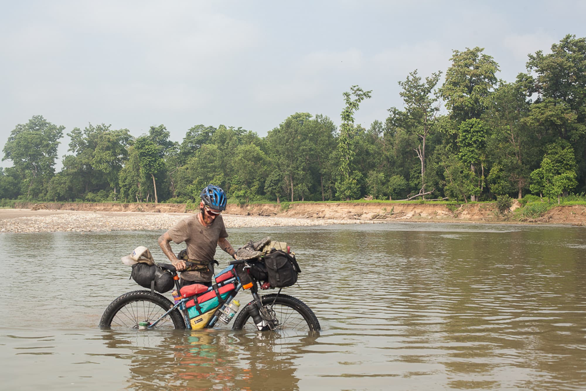 A cyclist rides a loaded bike across a shallow river.