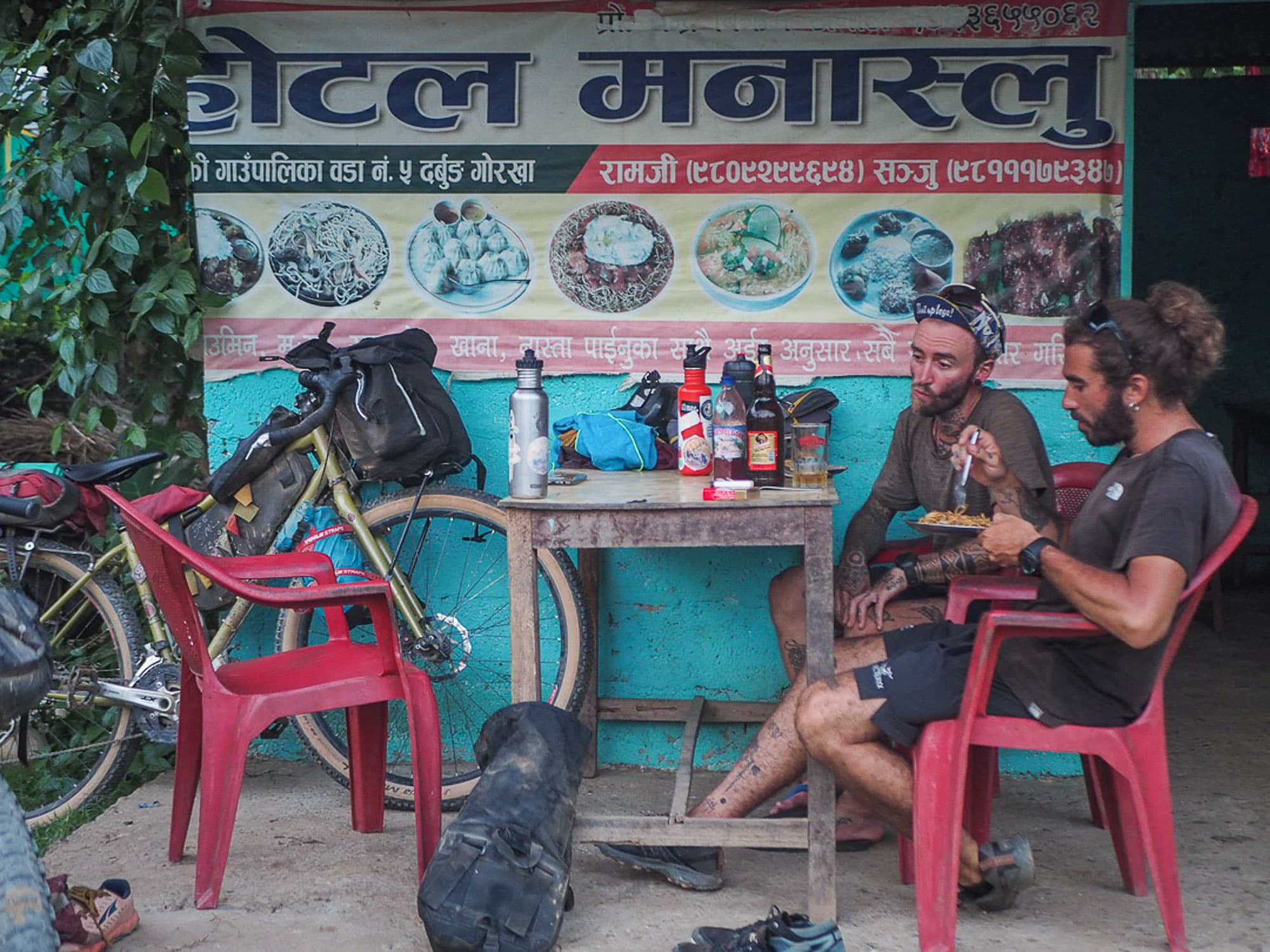 Two cyclists eat at a roadside cafe with their bikes leaned against the wall.