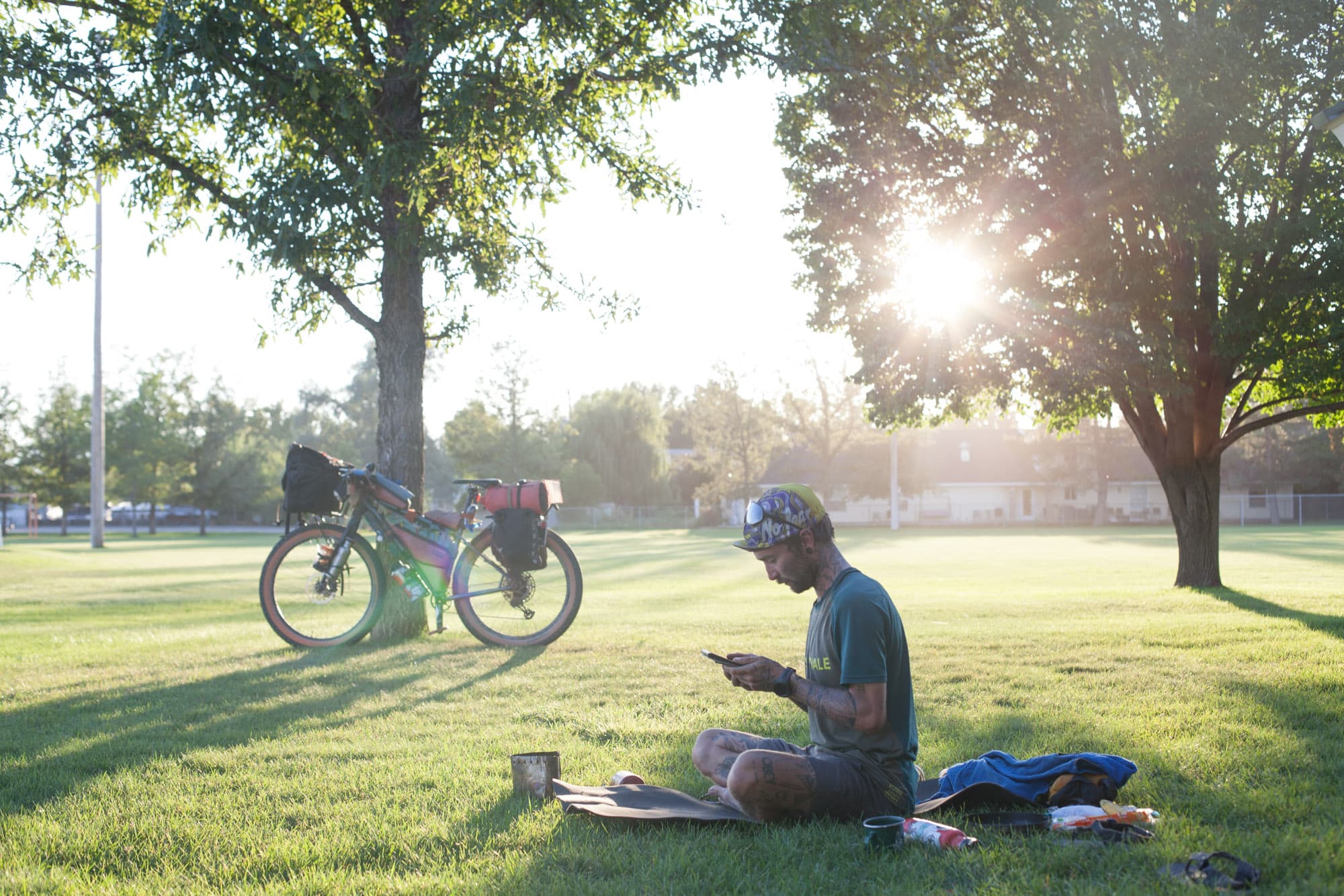 A loaded bike in a grassy park at golden hour as a rider sits and reads.