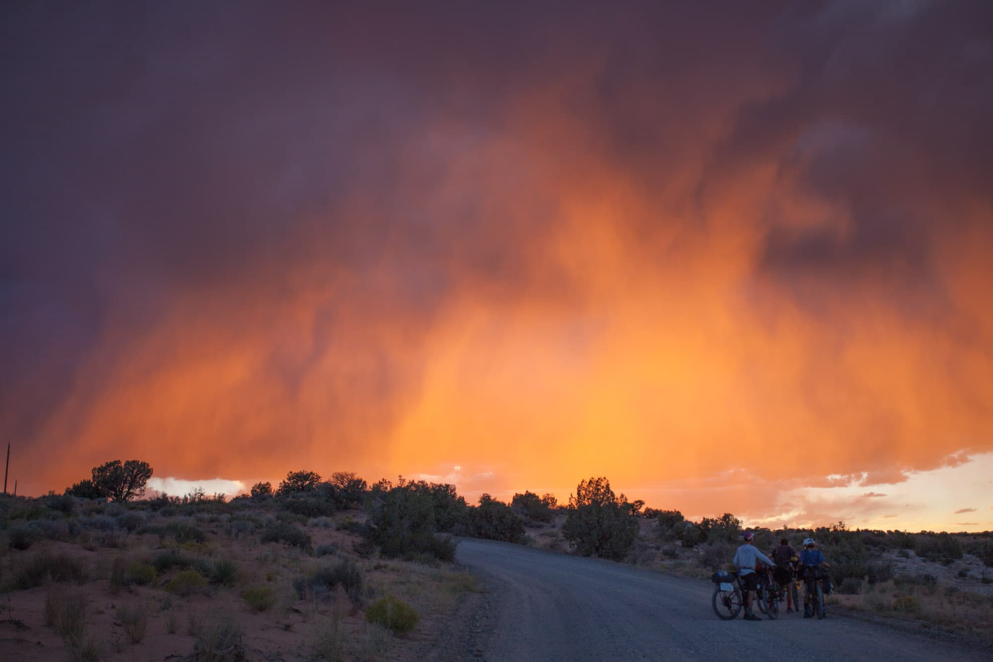 A dramatic orange storm sky over the desert with cyclists on a road below.