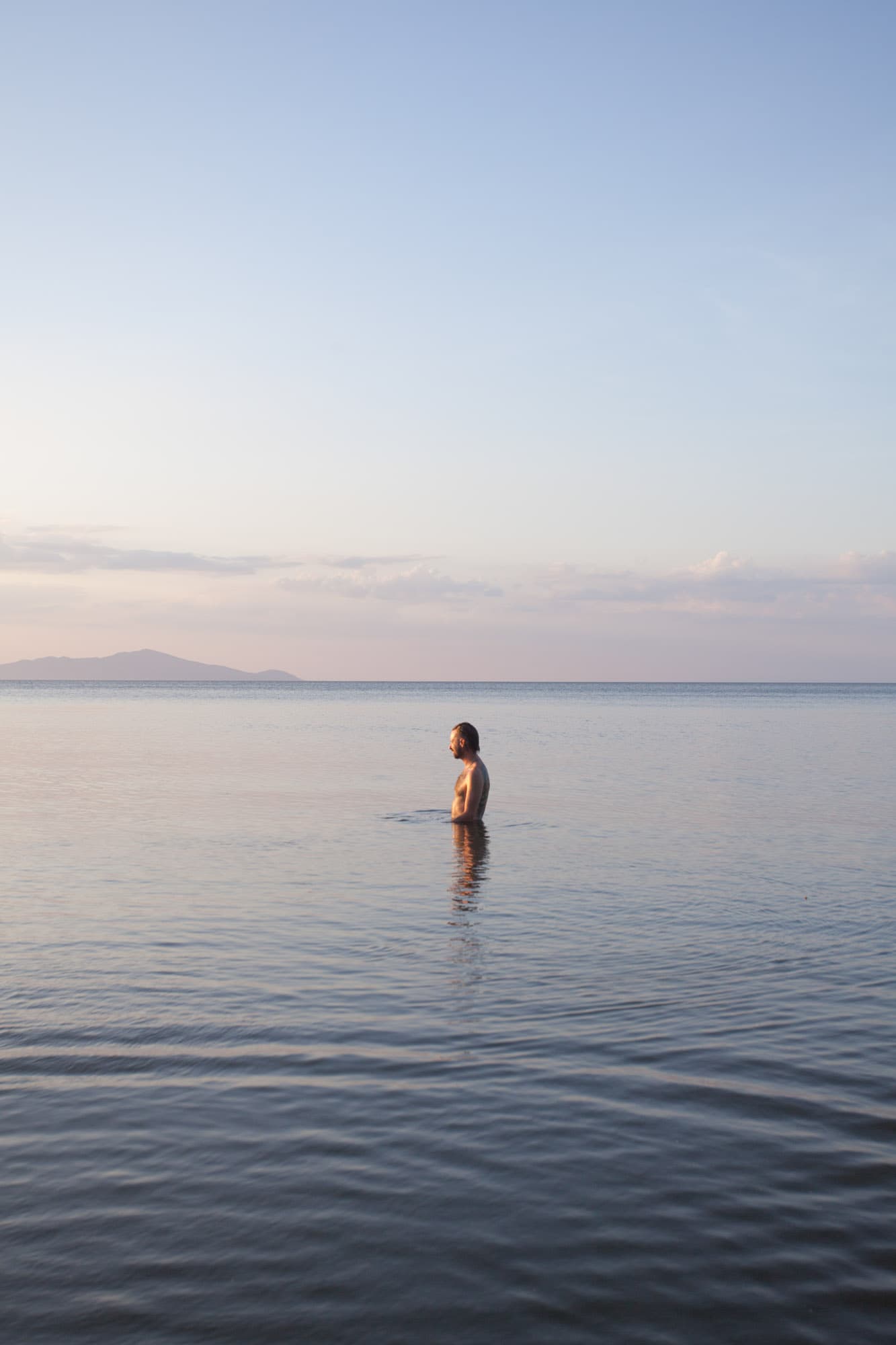 A figure stands waist-deep in a calm lake at dusk.