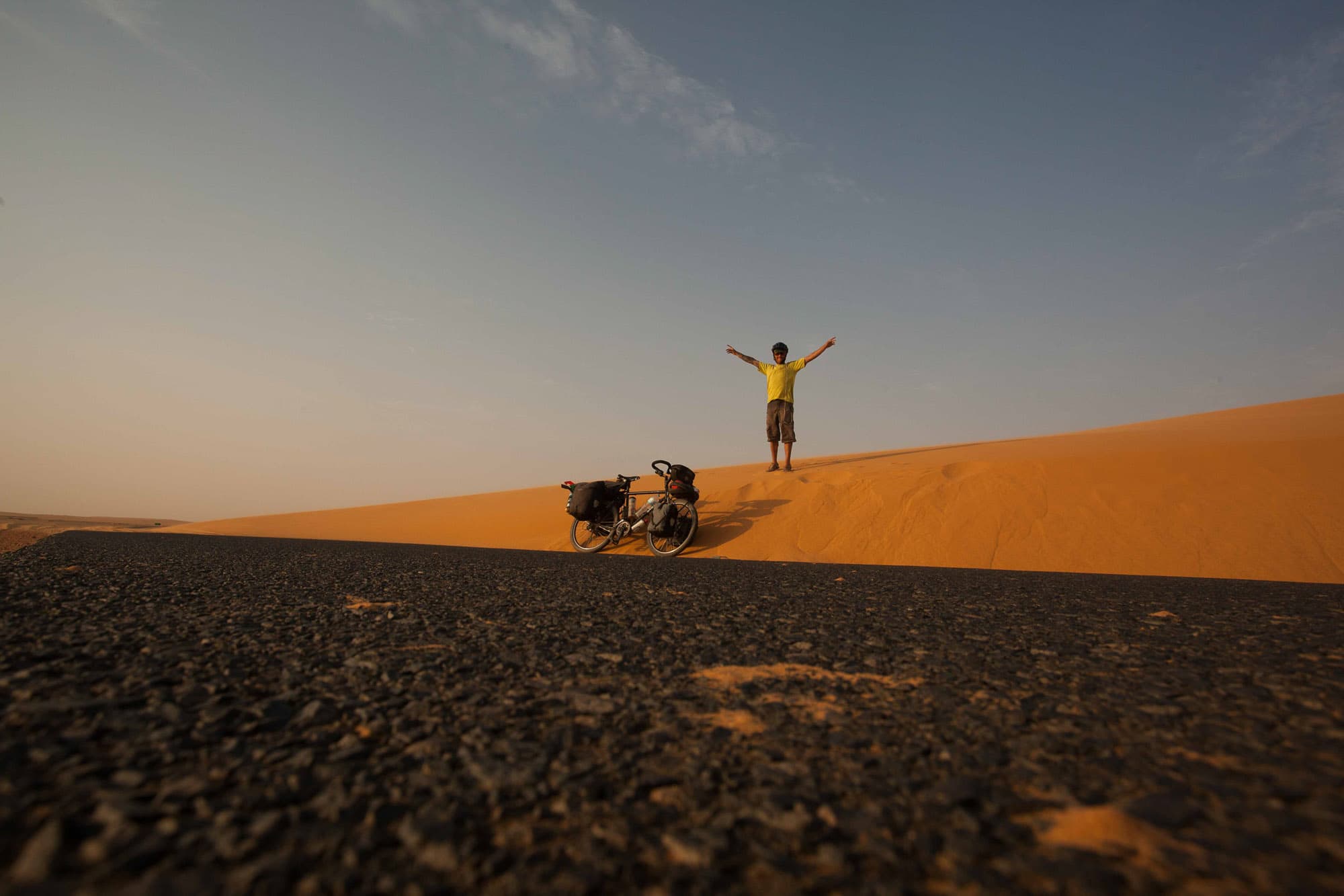 A cyclist stands with arms raised on an orange sand dune beside a loaded bike.