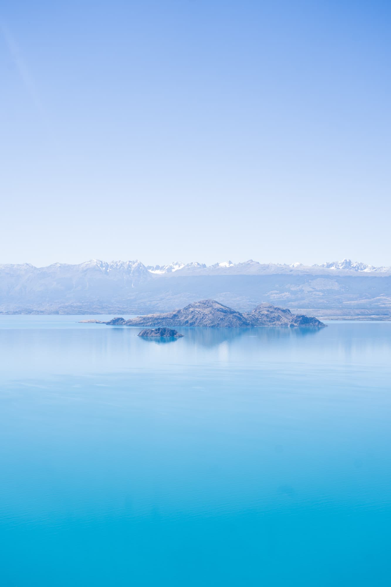 A turquoise lake with a small island and distant snow-capped peaks.
