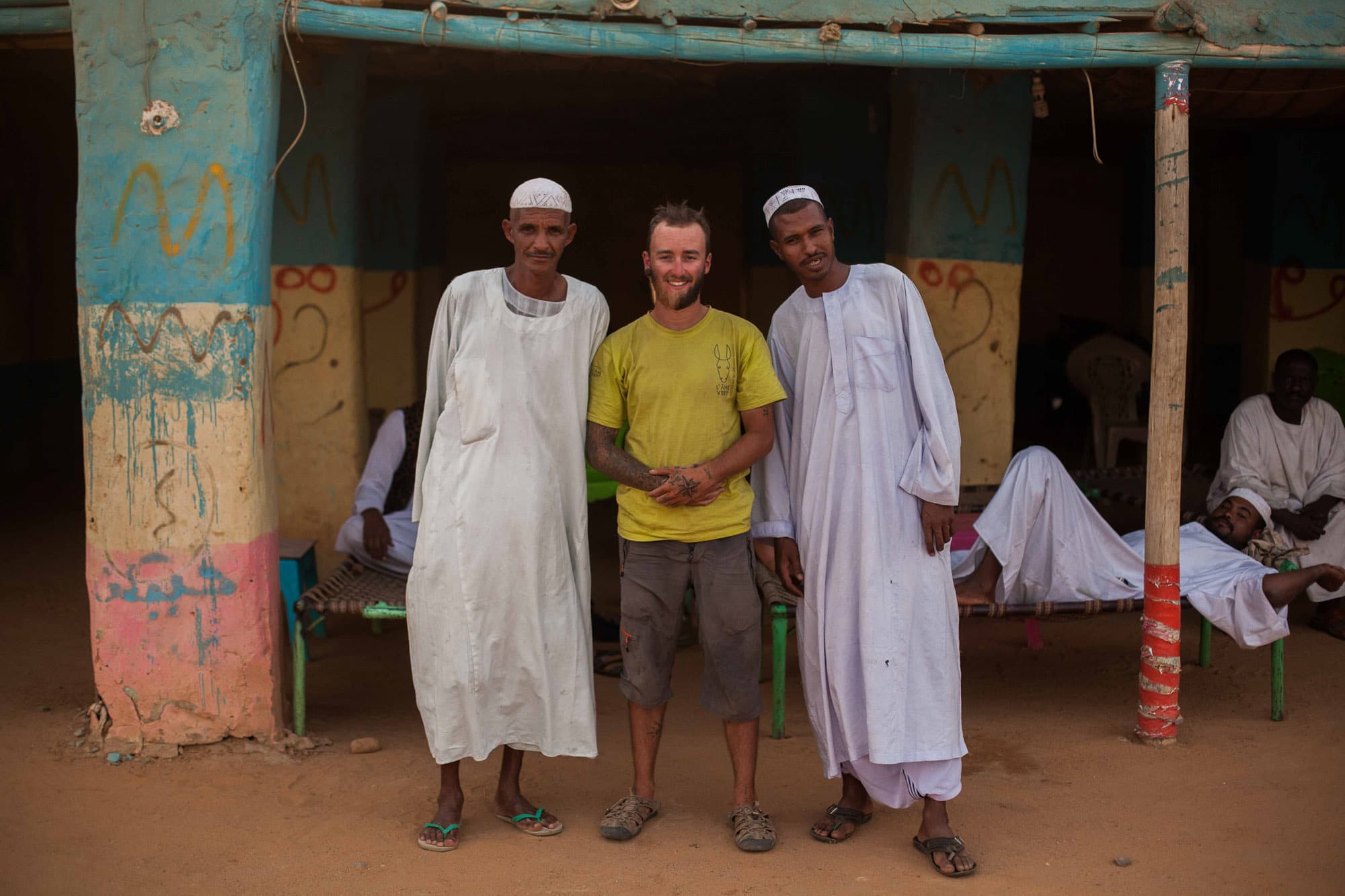 Clotaire stands between two men in white robes outside a painted building.