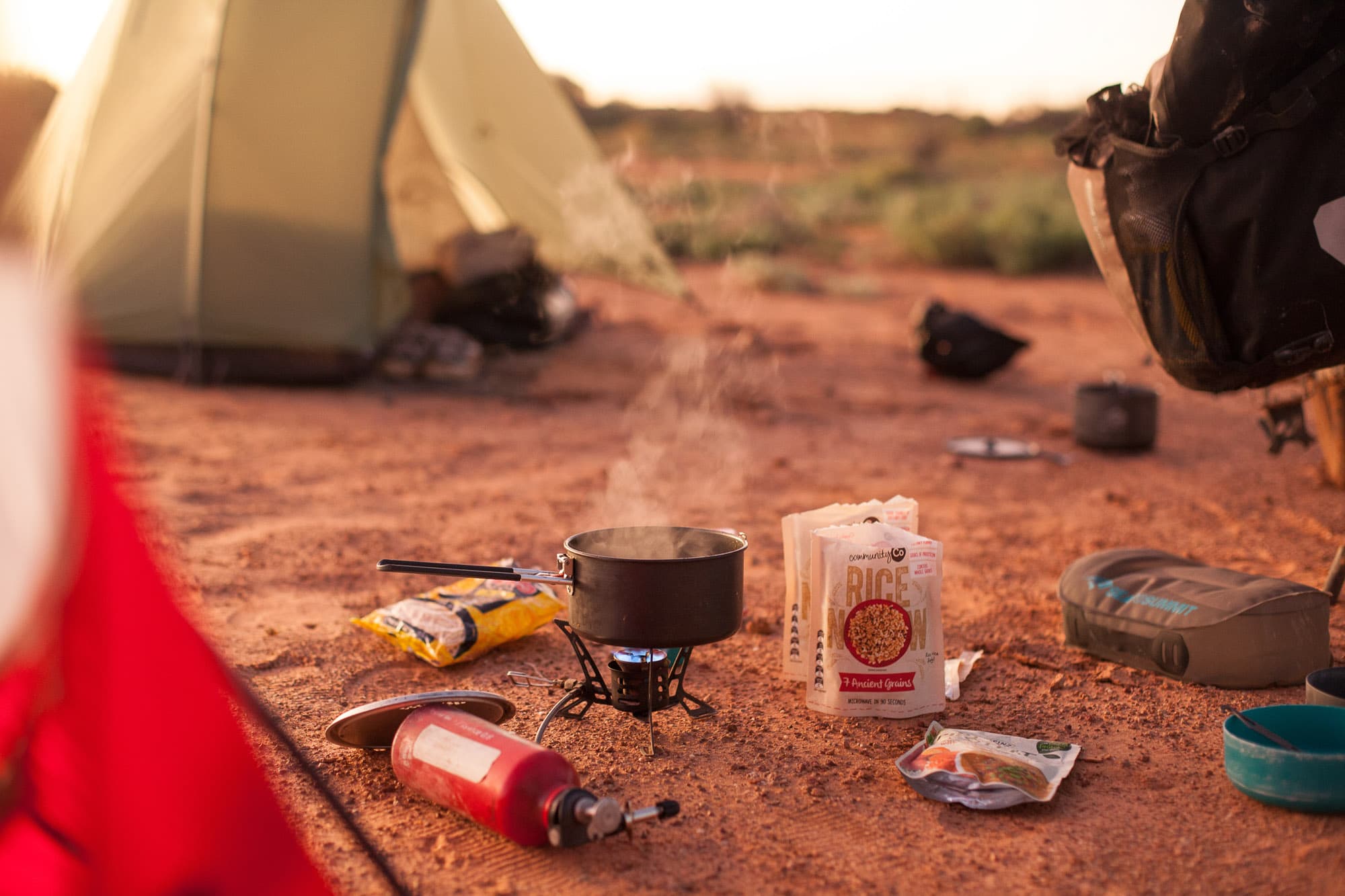 A camp stove and supplies on red desert sand beside a tent.