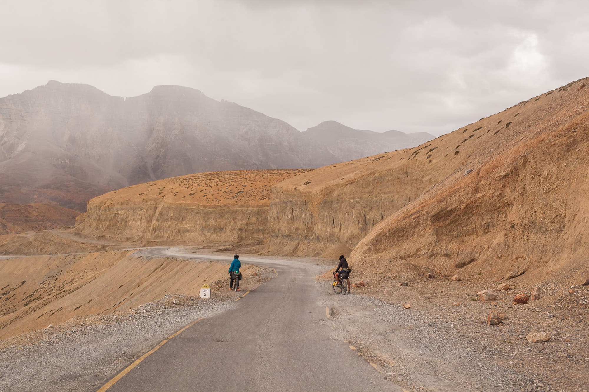 Two cyclists ride a paved road through arid mountains.