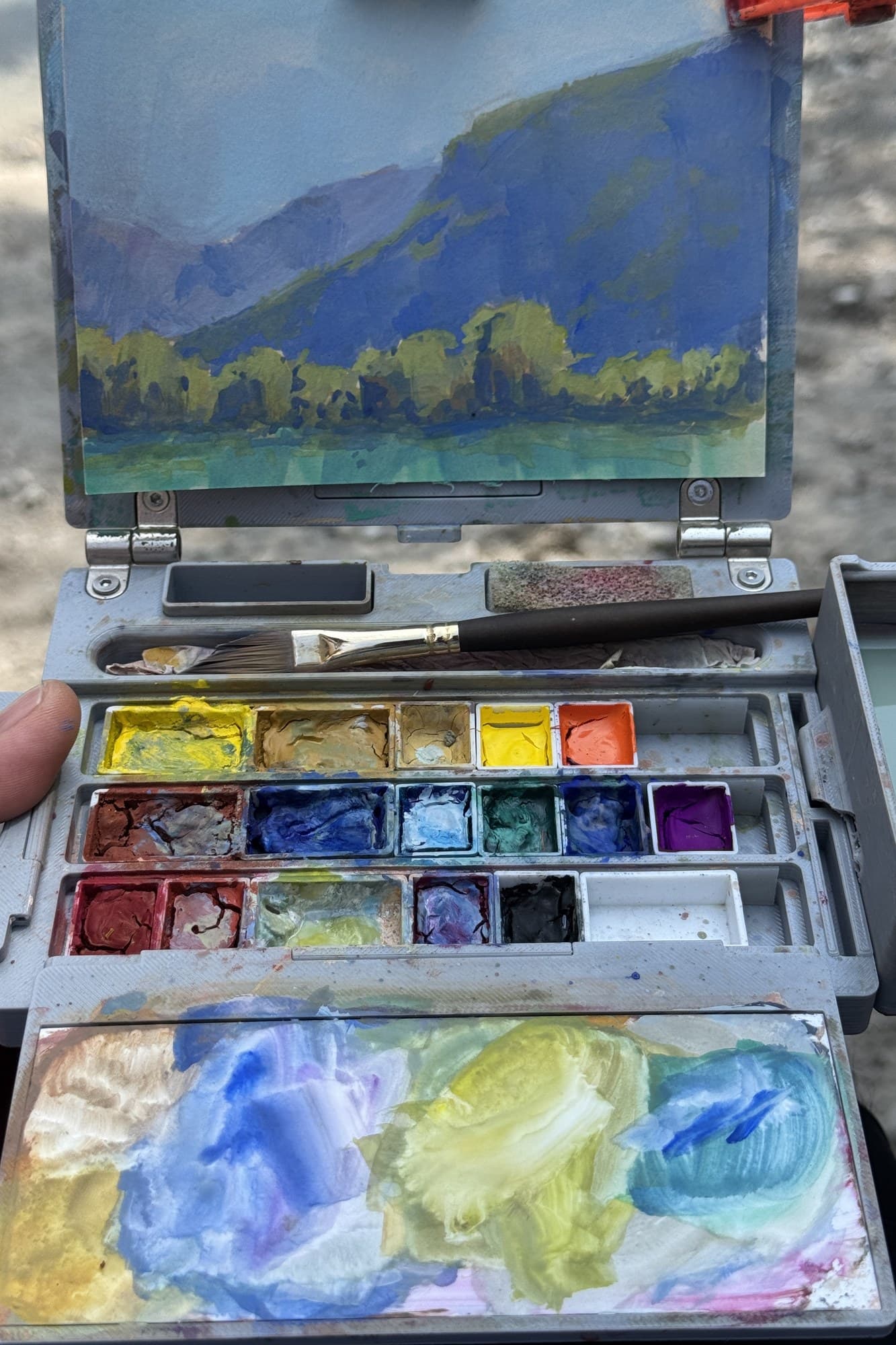 Open watercolor kit with a mountain painting clipped to the lid