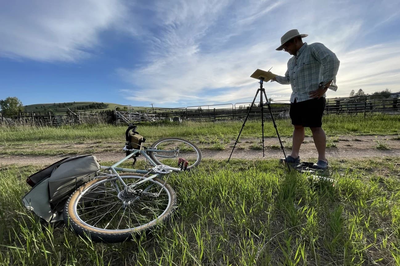 Russ painting at a tripod in a field with his bike lying in the grass