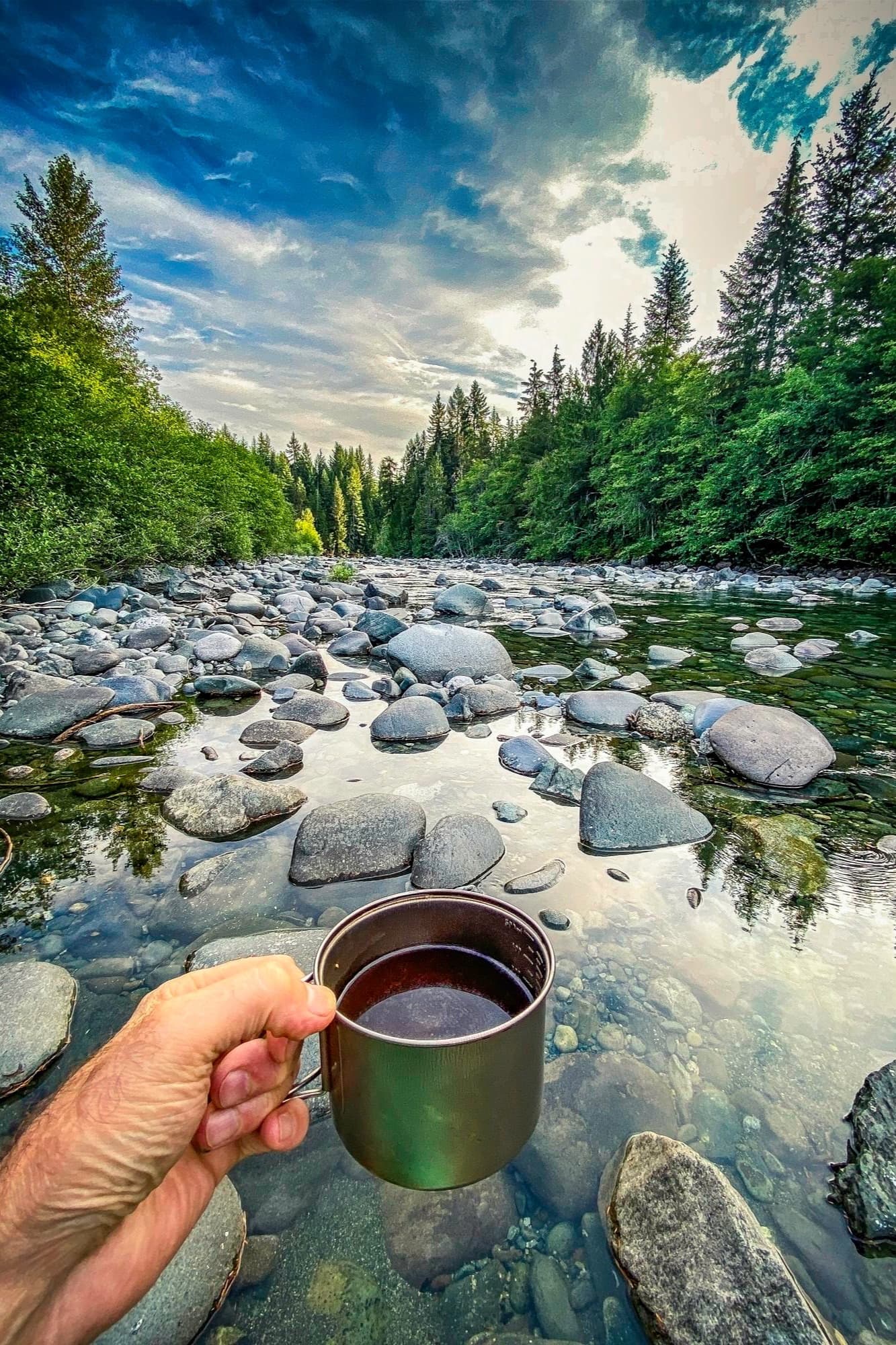 Morning coffee held over a clear rocky river