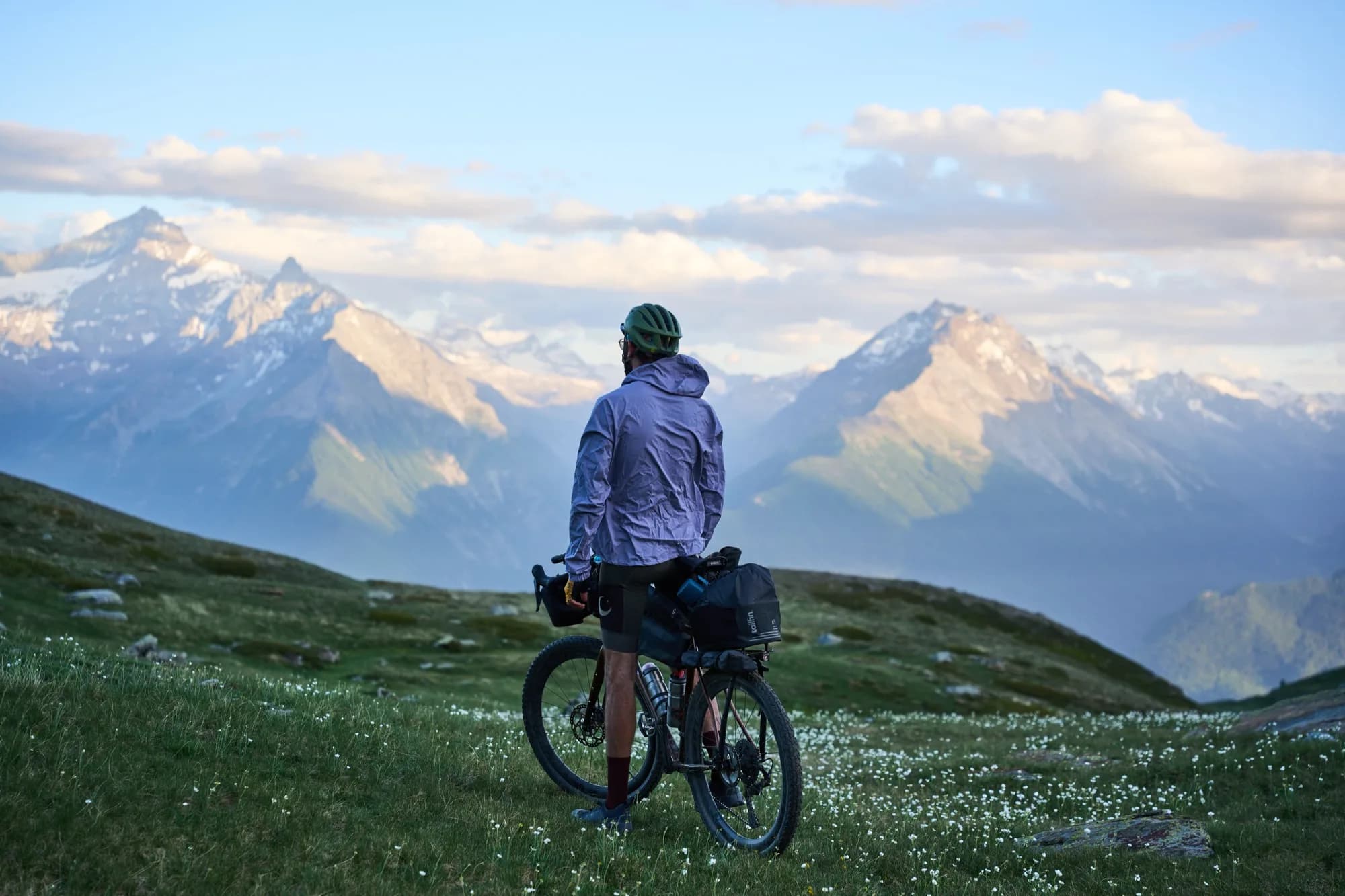 Davide with his bike in a flowering meadow at golden hour, facing the Alps