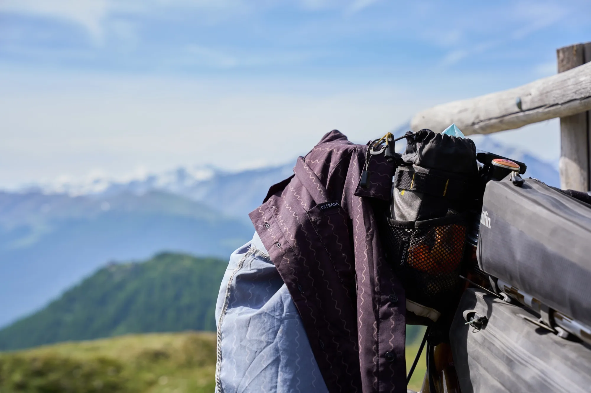 The Cascada shirt draped over the handlebar bags on a fence rail with the valley beyond