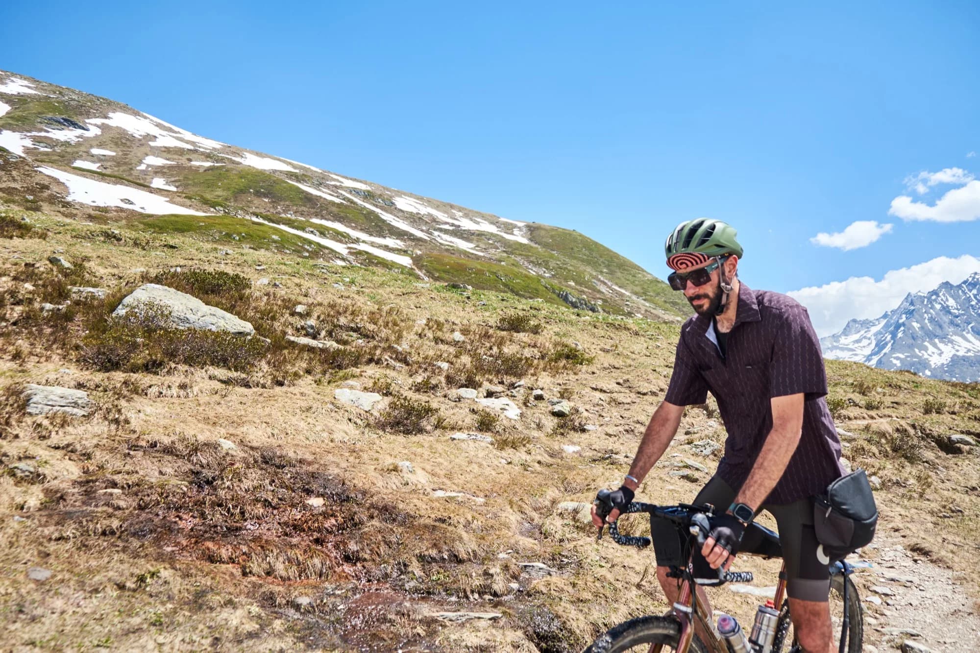 Davide riding a rocky gravel path toward the camera in the Cascada shirt with snowy peaks behind
