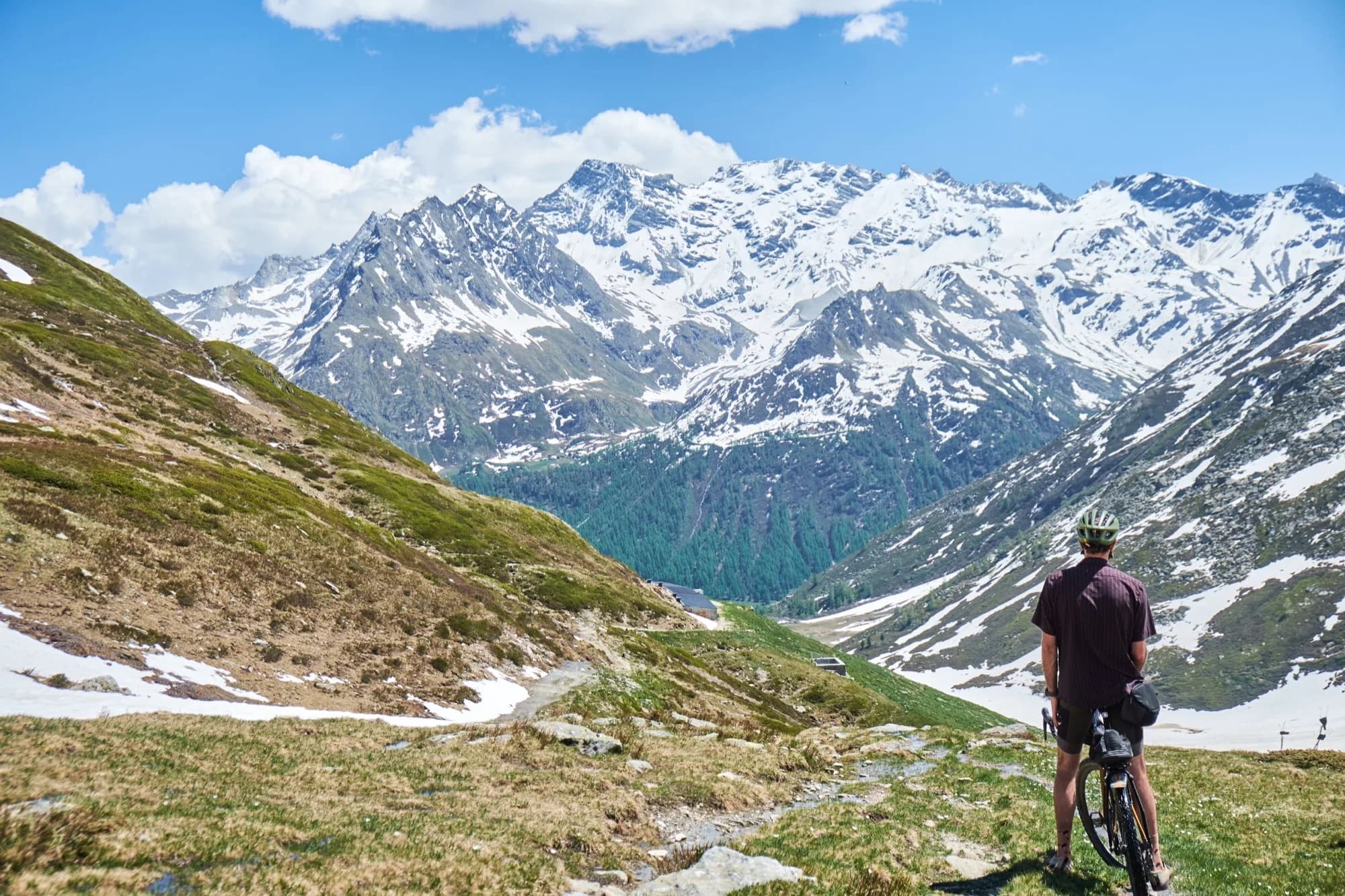Davide pausing with his loaded bike above a green Alpine valley ringed by snowy peaks