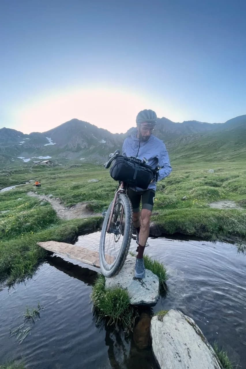 Davide carrying his bike across stepping stones over an alpine stream at dawn