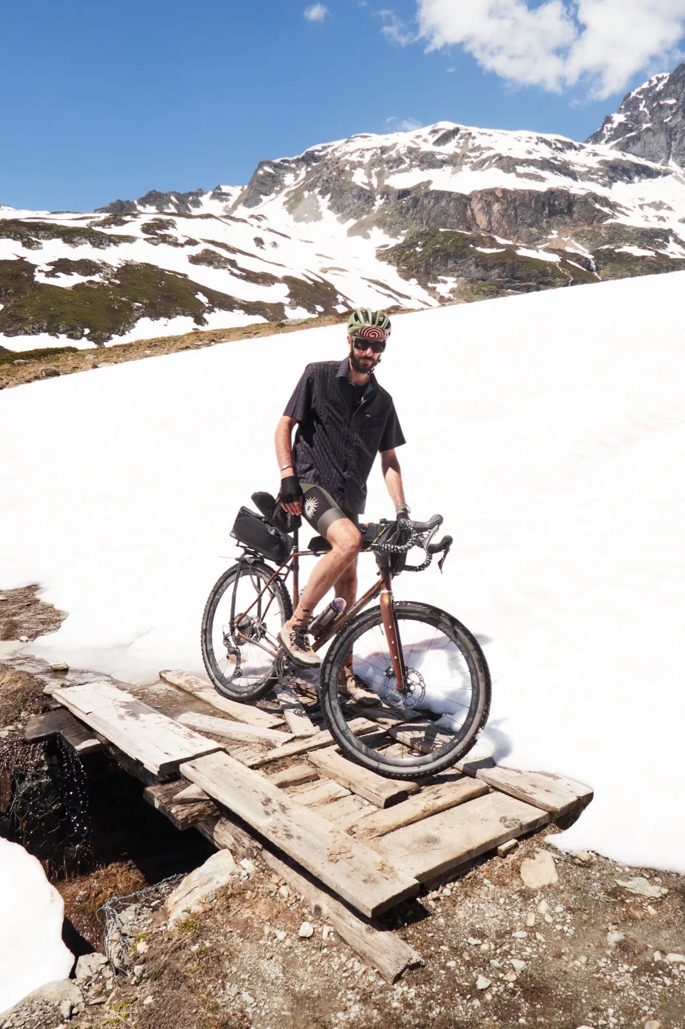 Davide on his loaded bike crossing a wooden plank bridge over snowmelt below a snowy peak