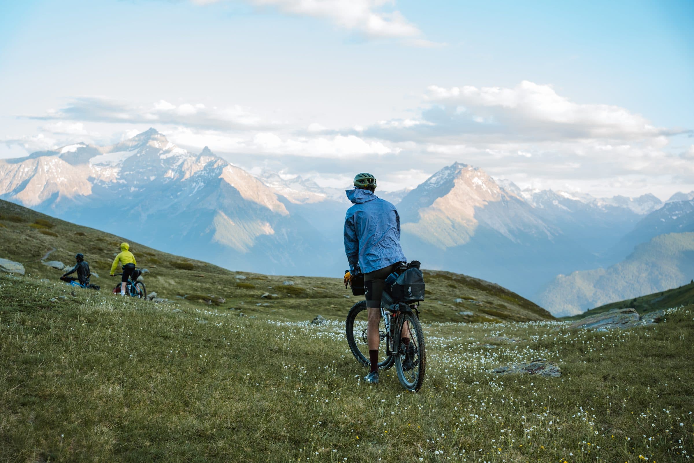 Bikepackers pausing in an Alpine meadow below snowy peaks.