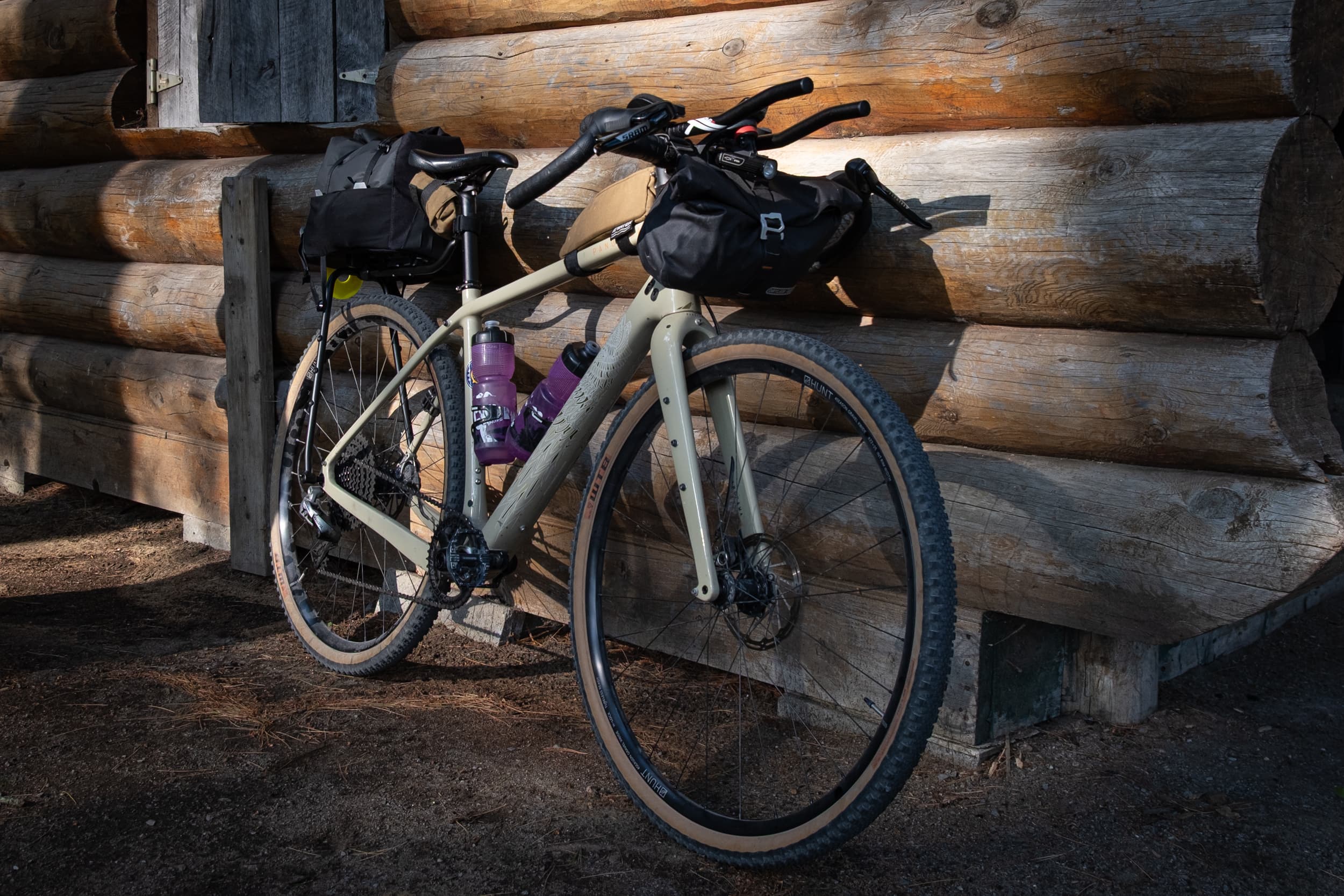 Loaded gravel bike with the Switch system against a log cabin wall in late-day light.
