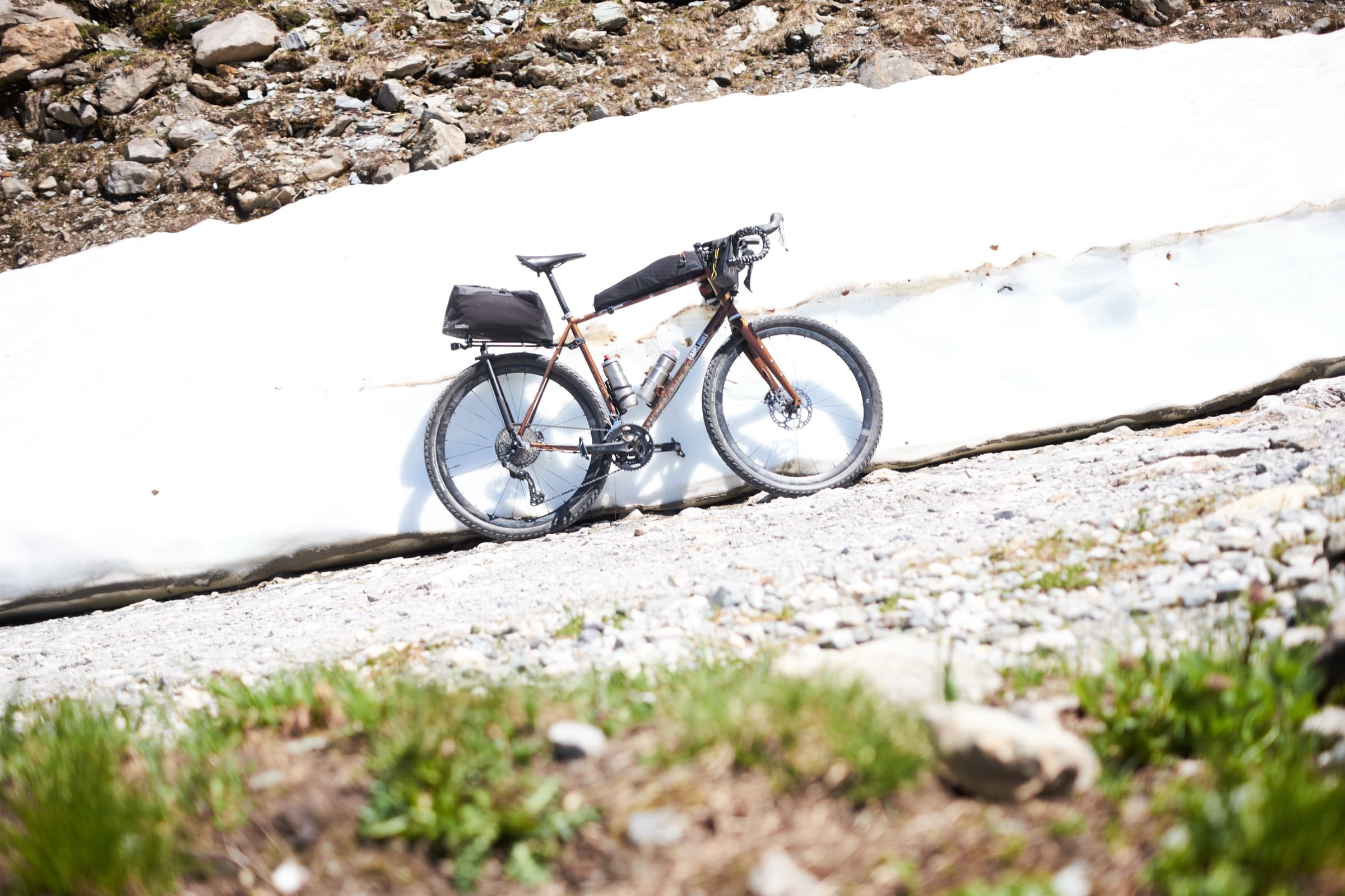 The bike parked beside a late-season snow patch on a gravel mountain road.