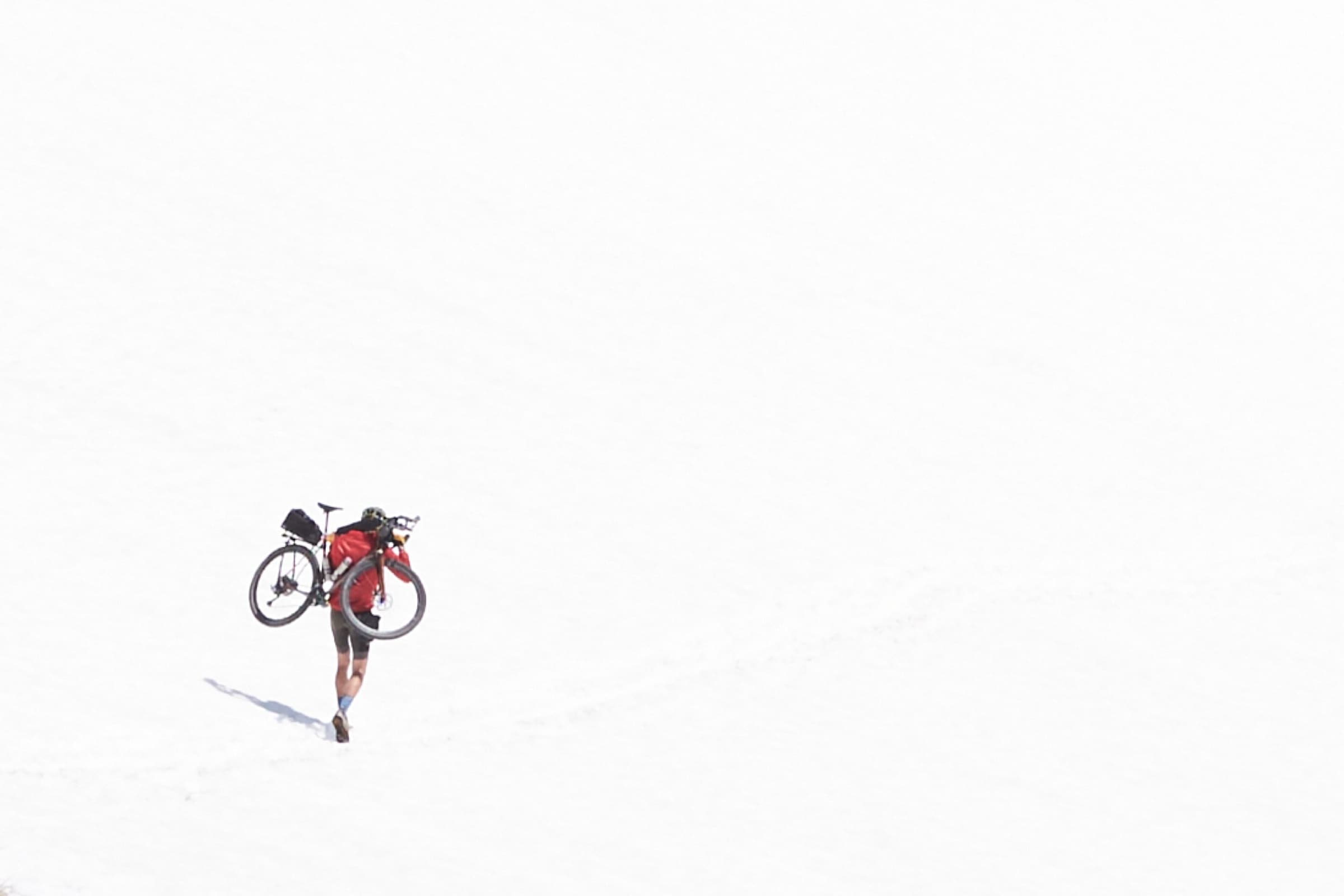 Davide carrying his loaded bike across a deep snow slope, a tiny figure against a vast white field.