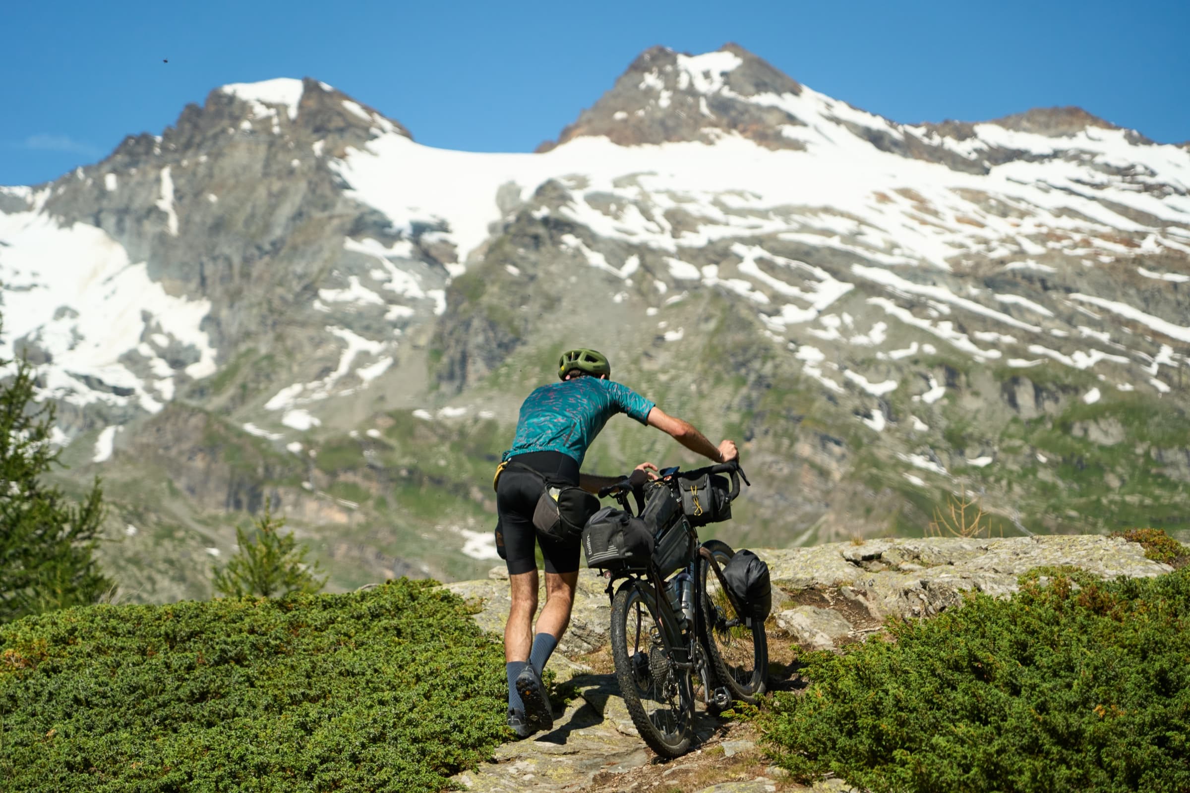 Davide pushing his loaded bike up a rocky alpine ridge, snow-capped peaks ahead.