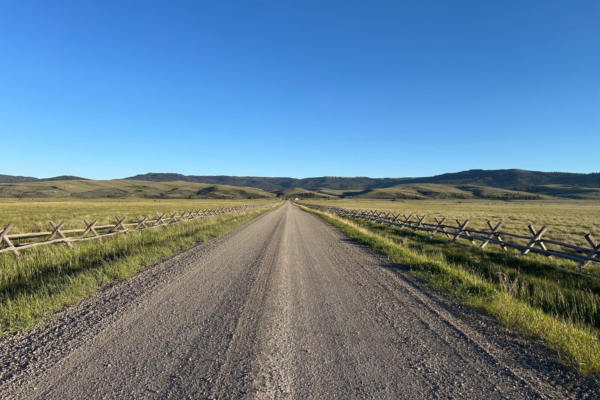 A straight gravel road lined with buck-and-rail fences under a huge blue sky