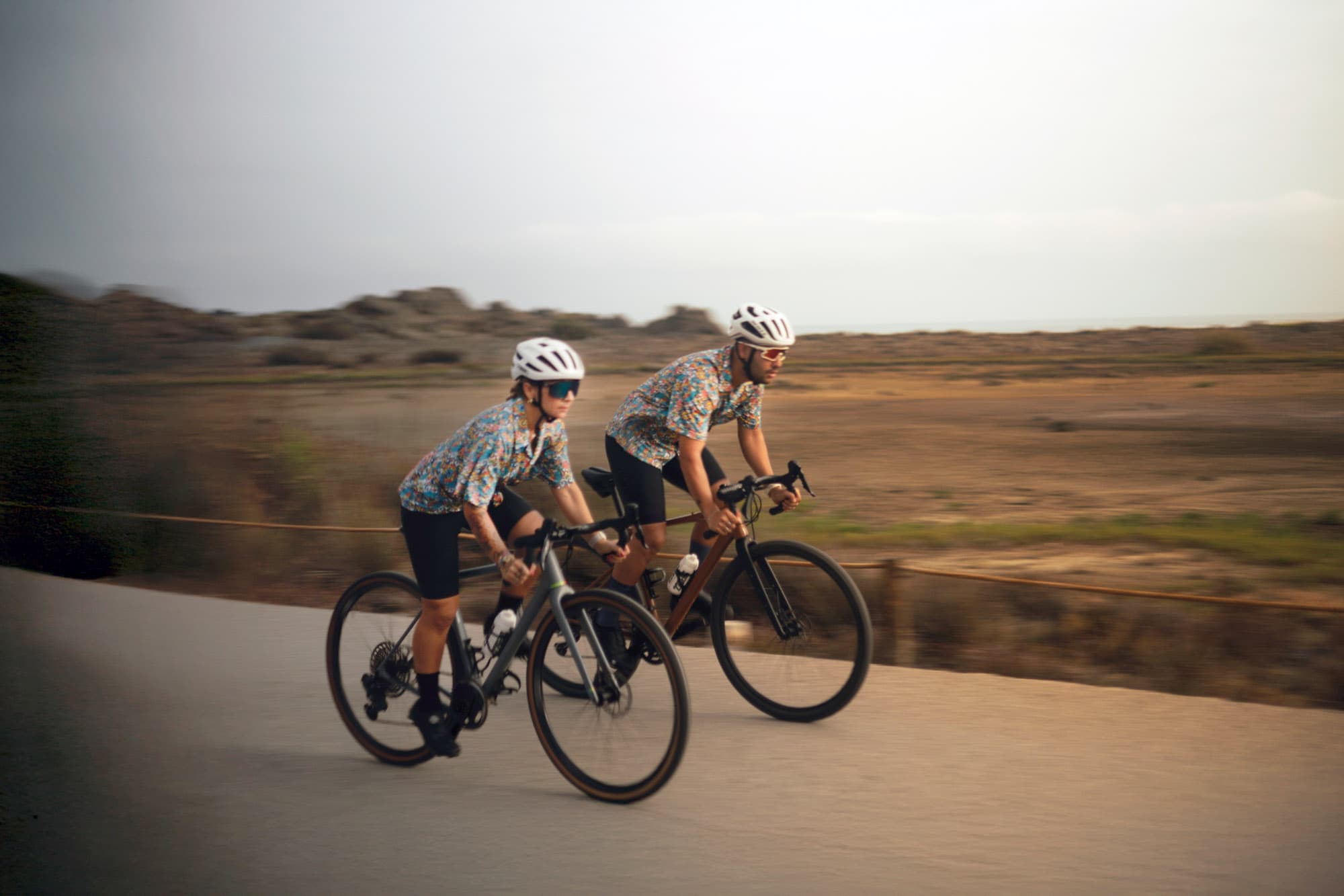 Two riders side by side in matching floral Pamera prints sprinting along a coastal road, motion blur.