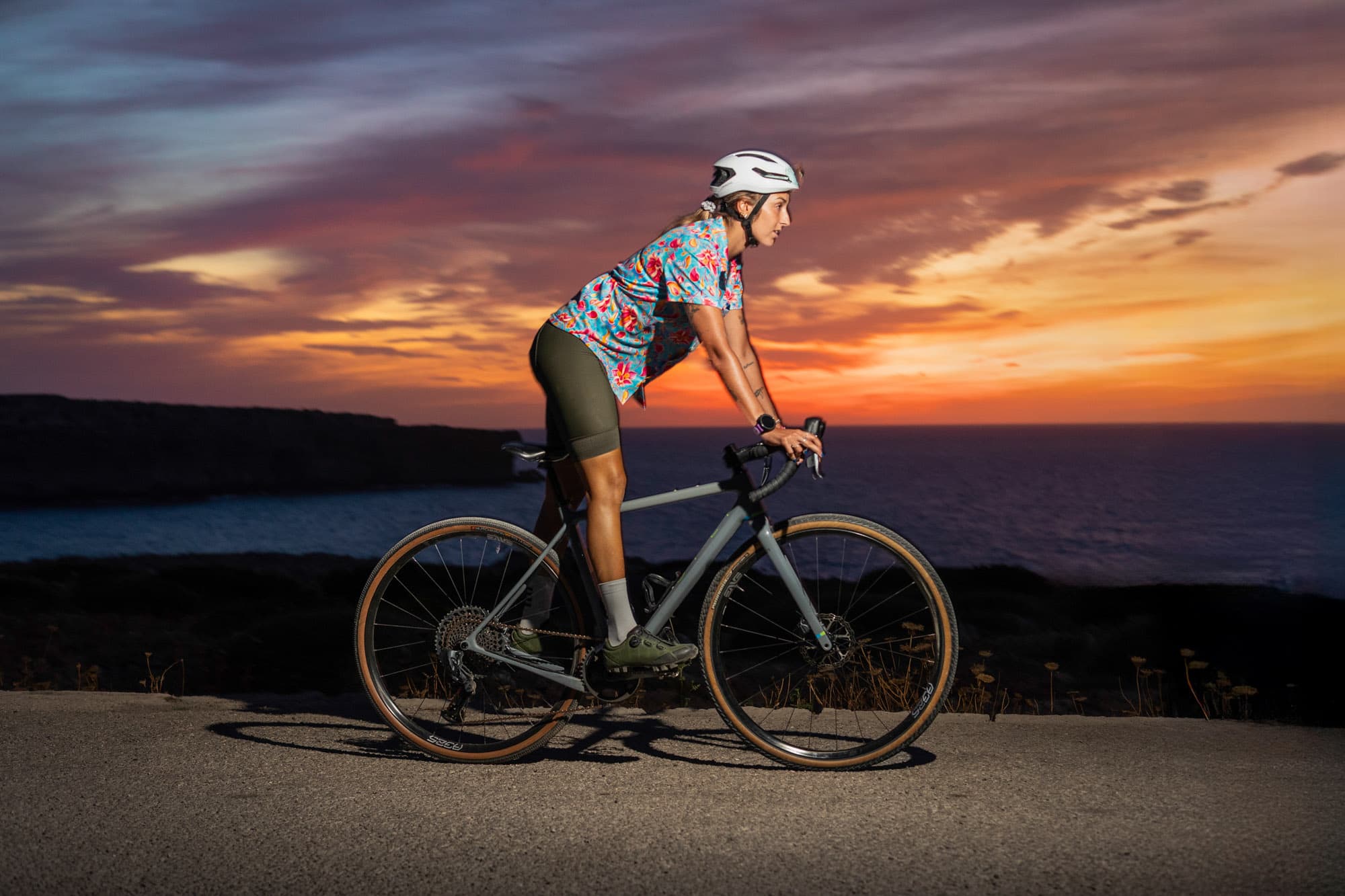 A solo Pamera rider paused on her gravel bike at sunset, the sea glowing orange behind her.