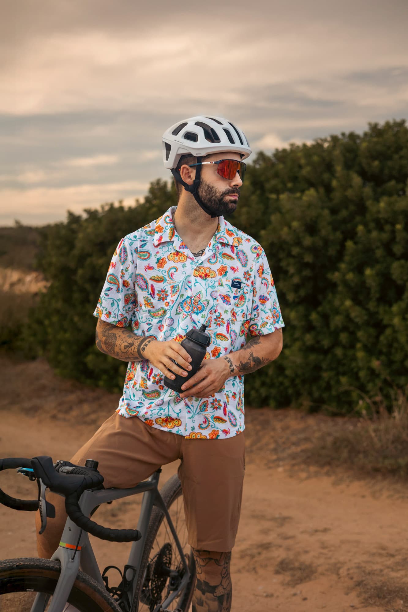 A male rider leaning on his gravel bike with a bottle, in a colourful paisley Pamera shirt.