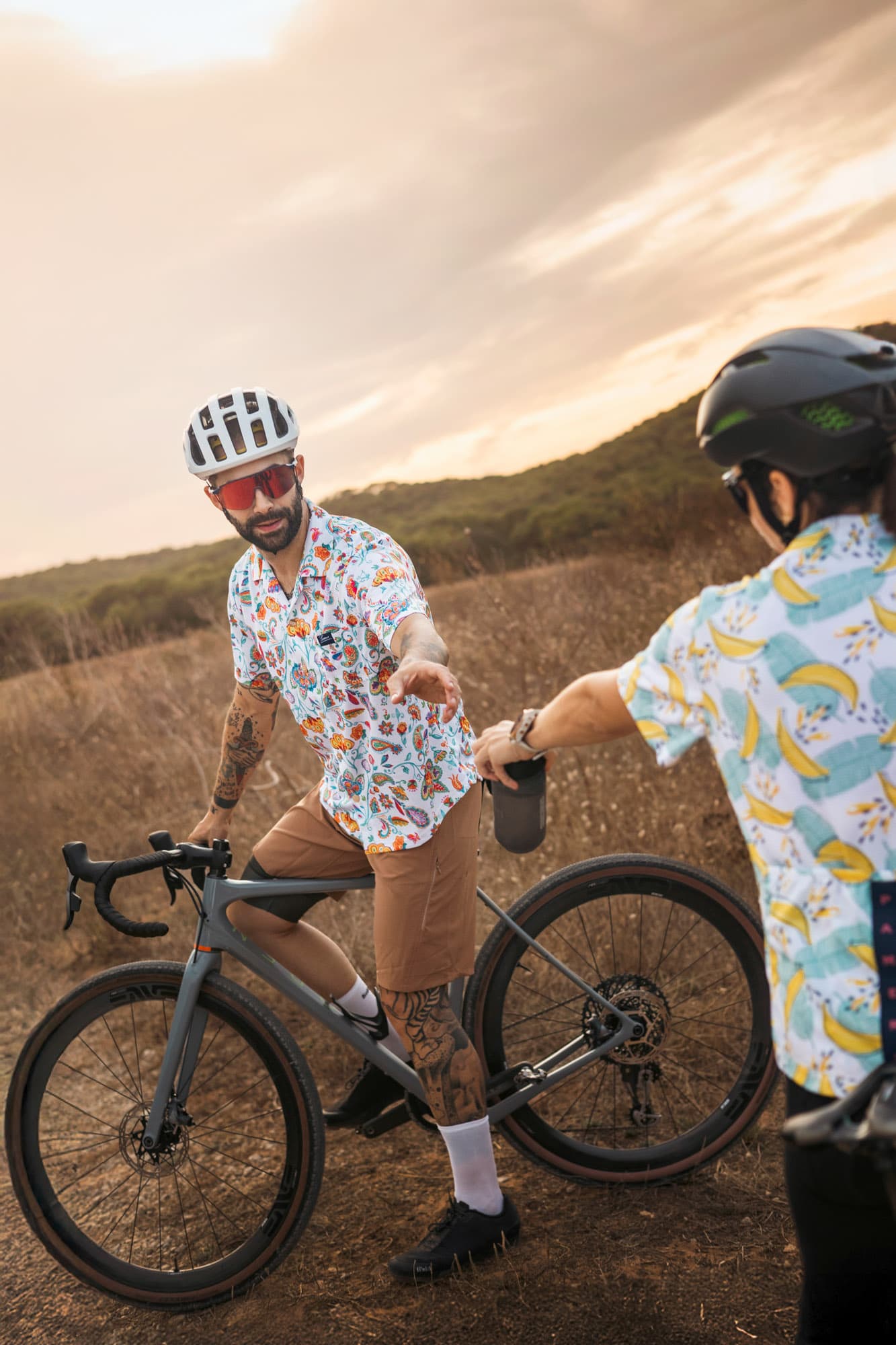 Two male riders pause at sunset to hand off a bottle, one in paisley print and one in banana print.