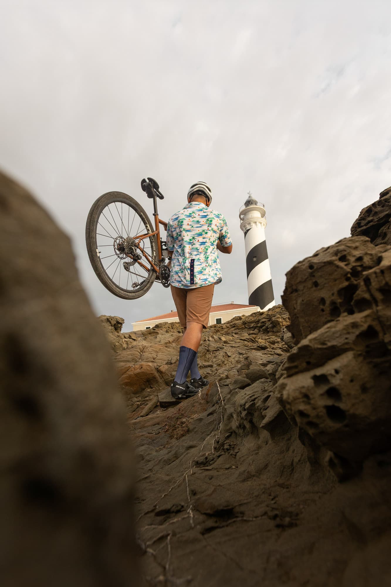 A rider hoists his gravel bike up rocks toward a black-and-white striped lighthouse, in a teal Pamera shirt.