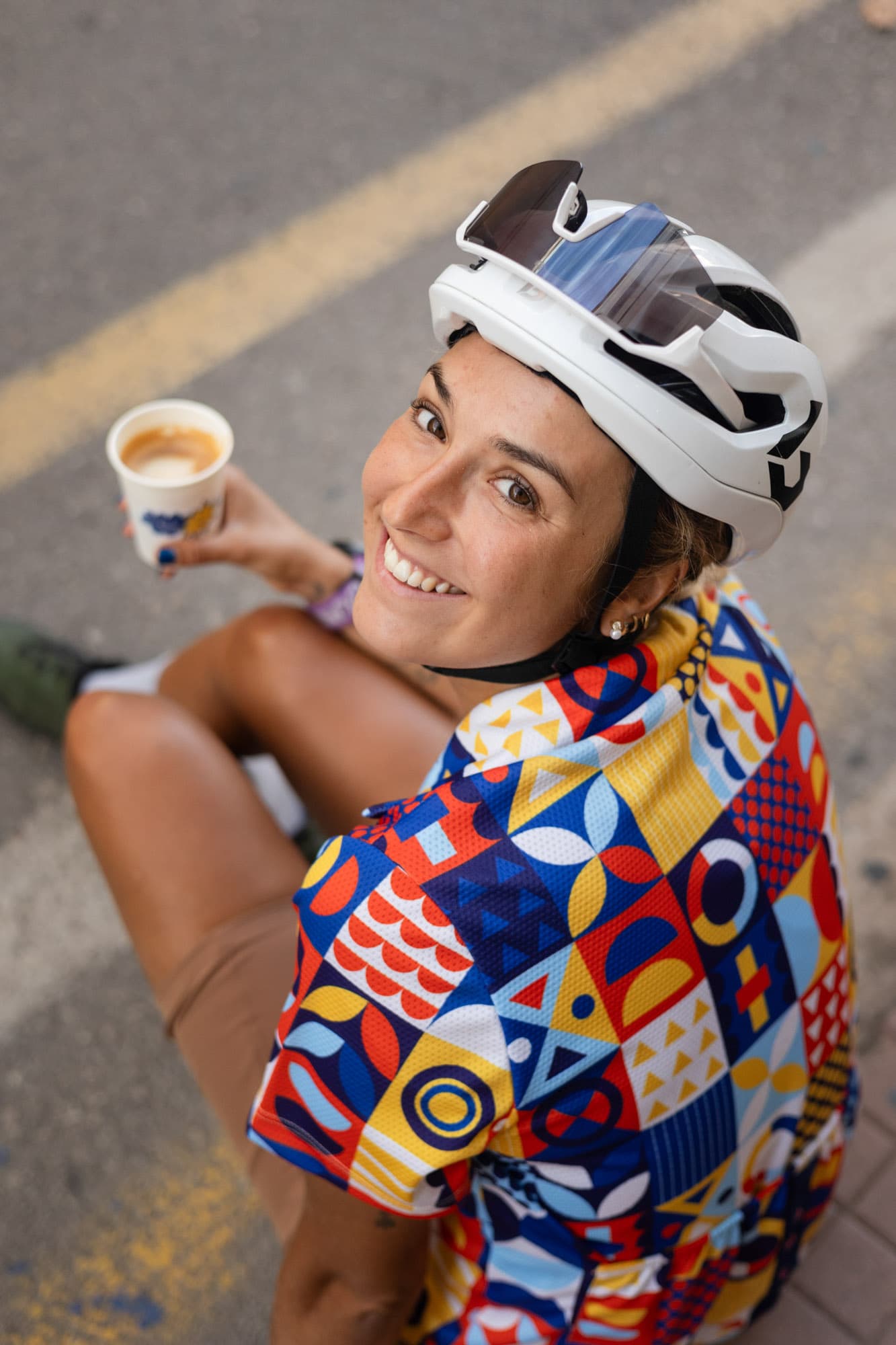 A smiling woman in a bold geometric Pamera shirt holds a small espresso cup at a coffee stop.