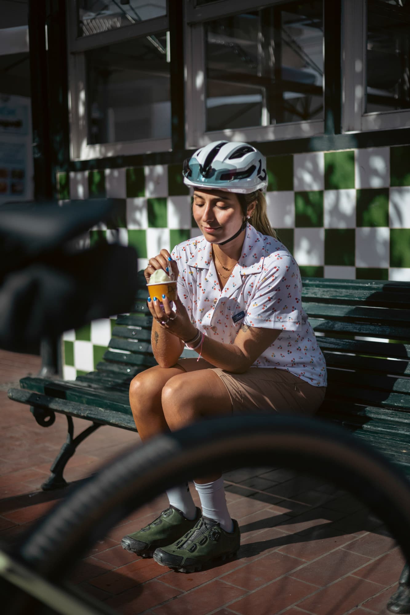 A woman in a cherry-print Pamera shirt eats gelato on a green bench, bike tyre in the foreground.