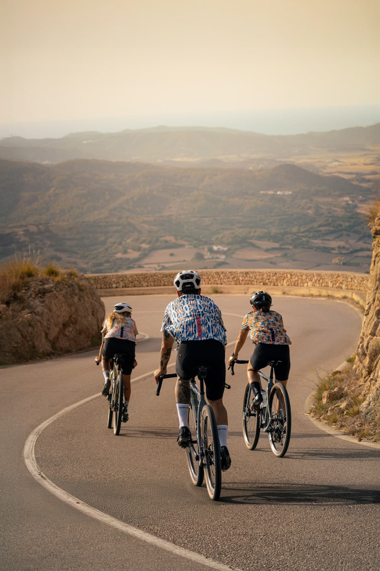 Three riders from behind climbing a coastal road into a golden Mediterranean valley.