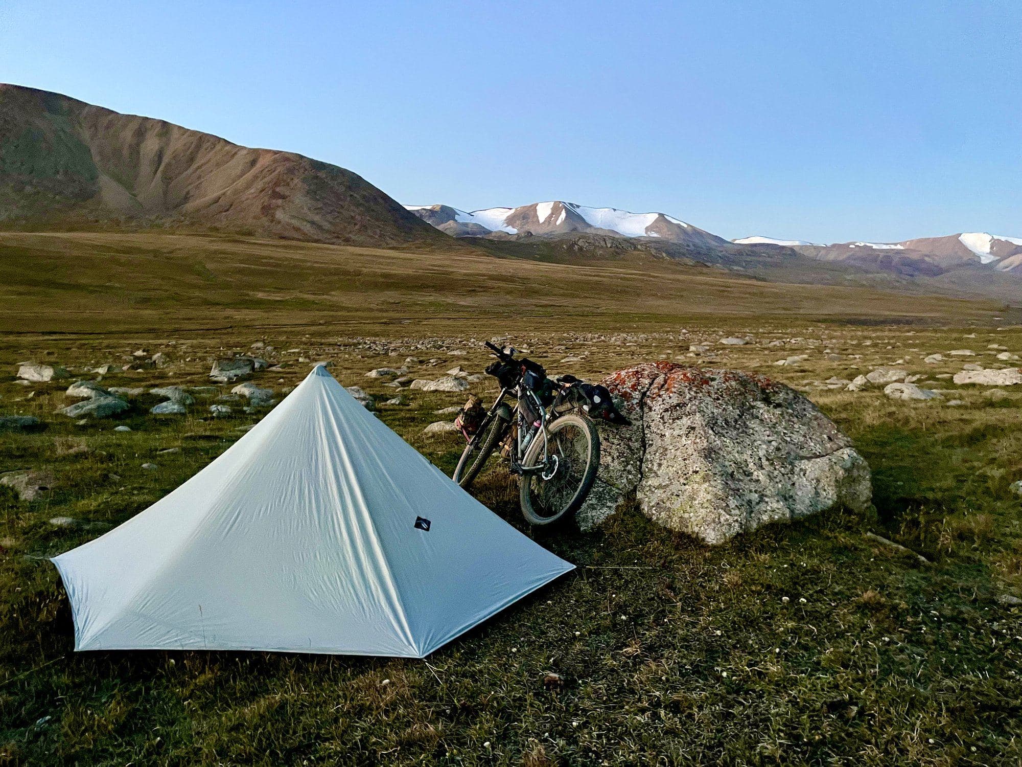A Six Moon Designs Lunar Solo tent pitched beside a boulder on a high alpine plateau, snowcapped peaks behind.