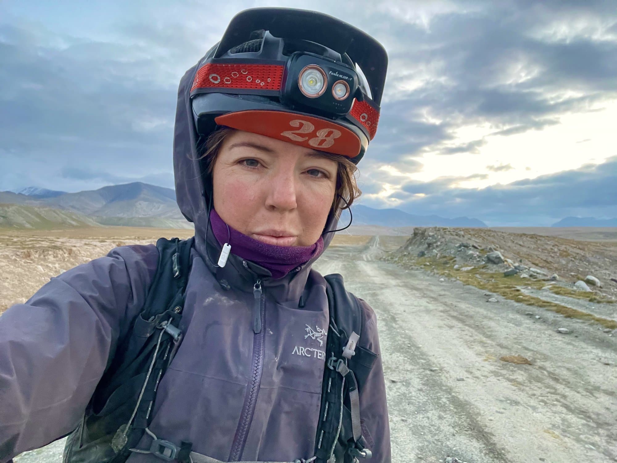 Britt in a rain shell and helmet with headlamp on a cold, open gravel road at altitude.