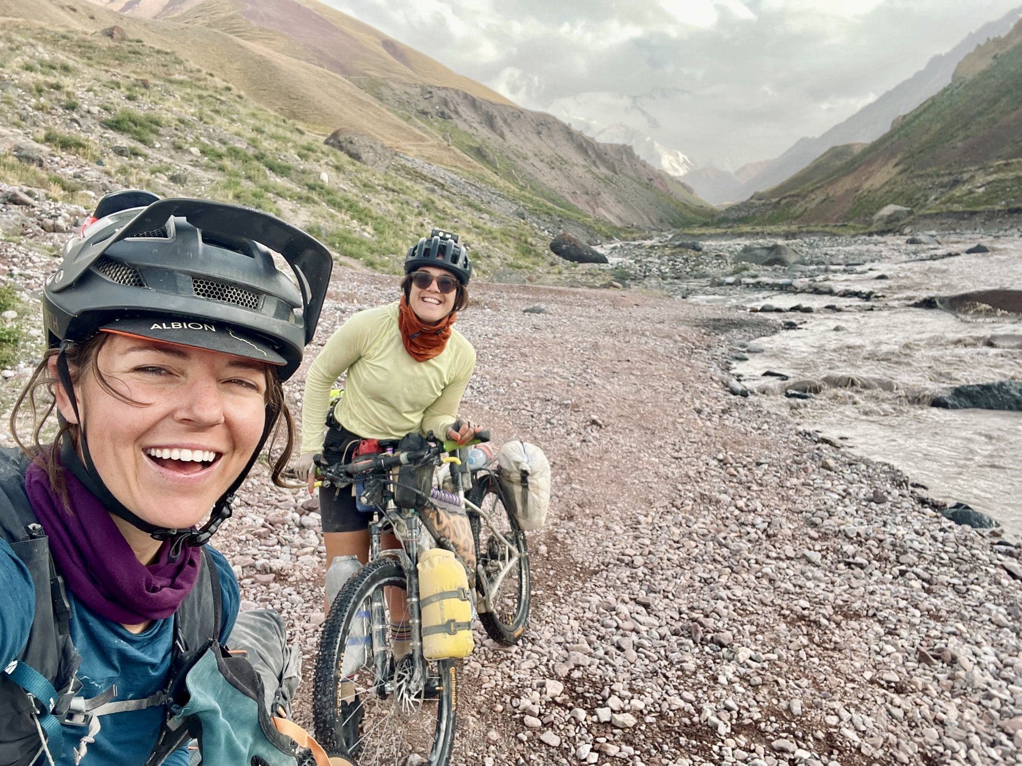 Britt grinning in a selfie with a fellow racer on a wide gravel riverbed after a river crossing.