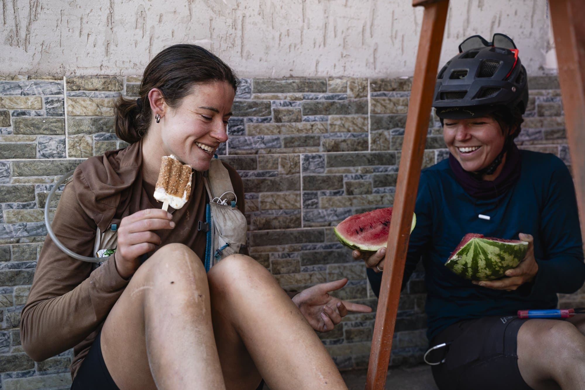 Two racers laughing over popsicles and watermelon during a roadside resupply.