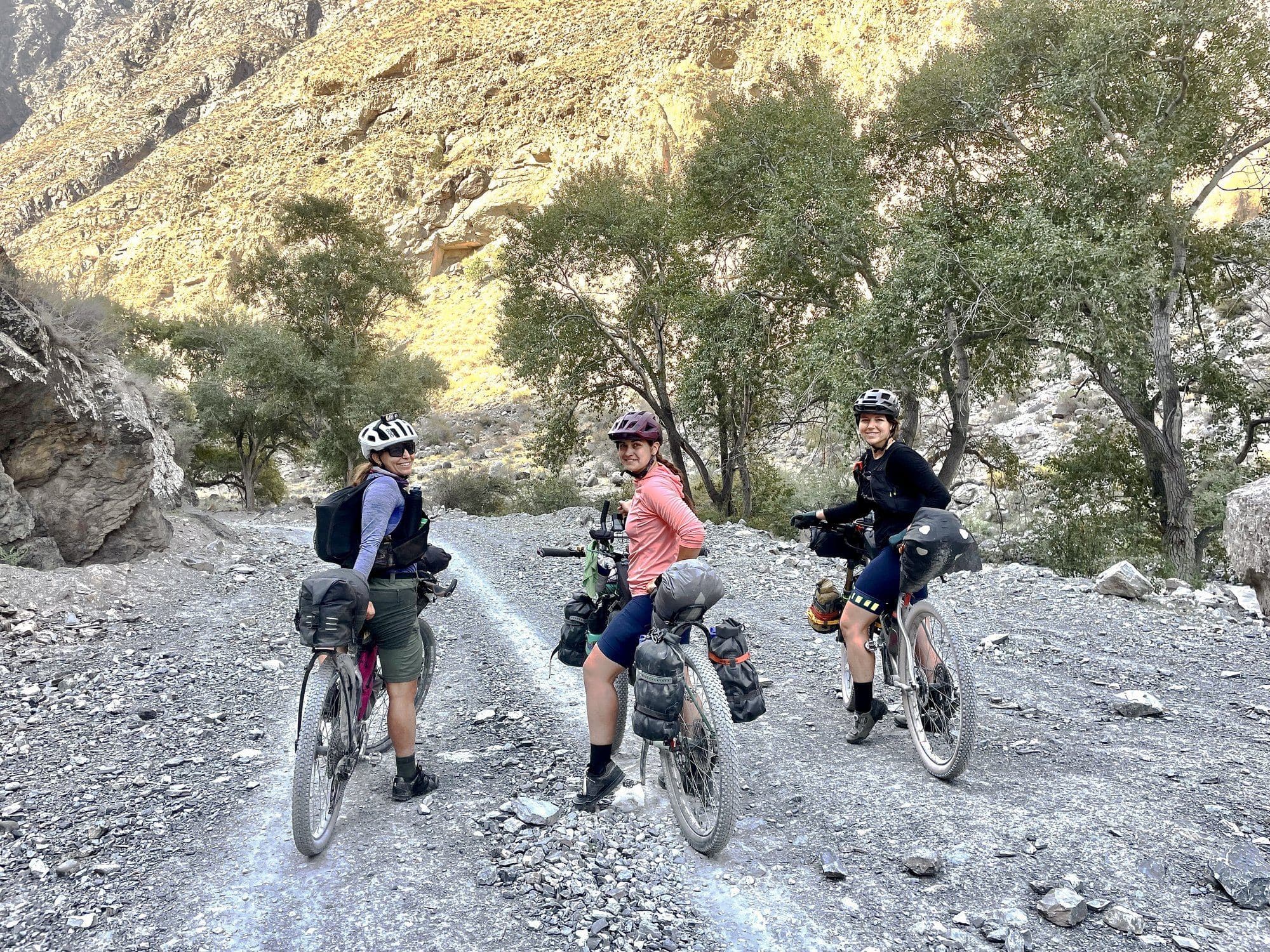 Three women racers stopped together on a rocky track below a sunlit canyon wall.