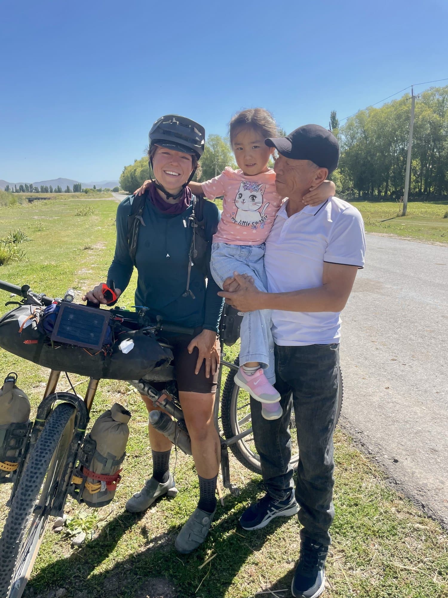 Britt with a local man holding a young girl, her loaded bike beside them on a country road.