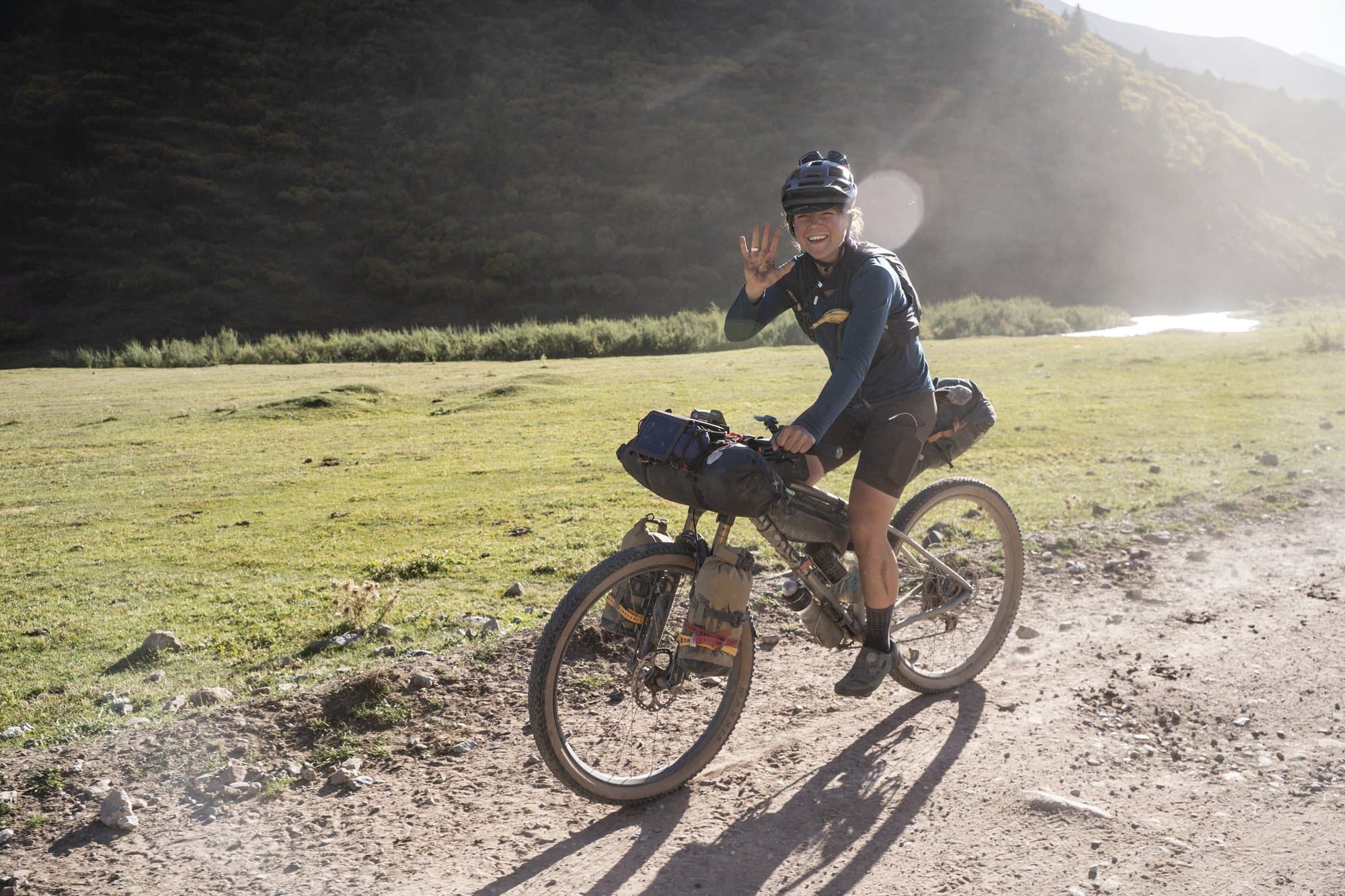 Britt waving from the saddle on a sunlit dirt track through a green meadow.