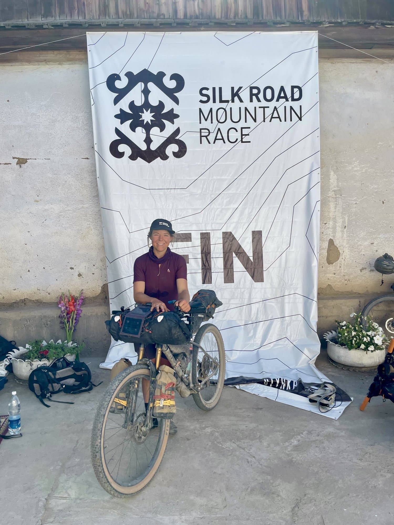 Britt sitting with her muddy bike under the Silk Road Mountain Race finish banner.